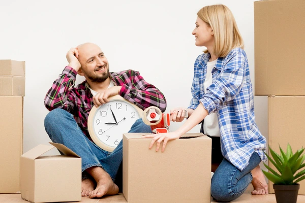 Zwei Personen in karierten Hemden sitzen zwischen Umzugskartons, eine hält eine Uhr, die andere klebt einen Karton ab.