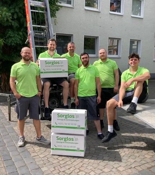 Sechs Umzugshelfer von Sorglos Umzüge & Entrümpelungen in grünen Poloshirts posieren vor einem Außenlift an einem Gebäude mit drei Kartons der Umzugsfirma