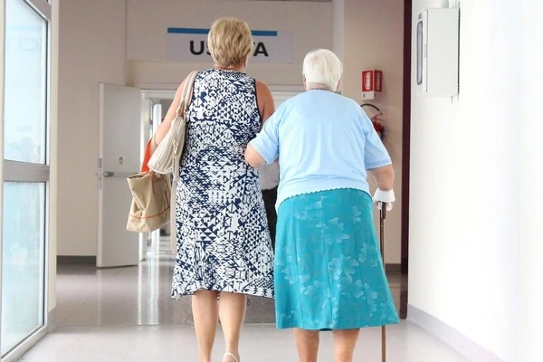 Zwei Frauen gehen gemeinsam einen hellen Flur eines Pflegeheims entlang. Die ältere Frau rechts stützt sich auf einen Gehstock und wird von der anderen Frau am Arm gehalten. Beide tragen Sommerkleidung mit Mustern.