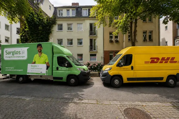 Das Sorglos-Mobil und ein DHL-Transporter auf einer gepflasterten Straße mit mehrere Reihenhäusern im Hintergrund